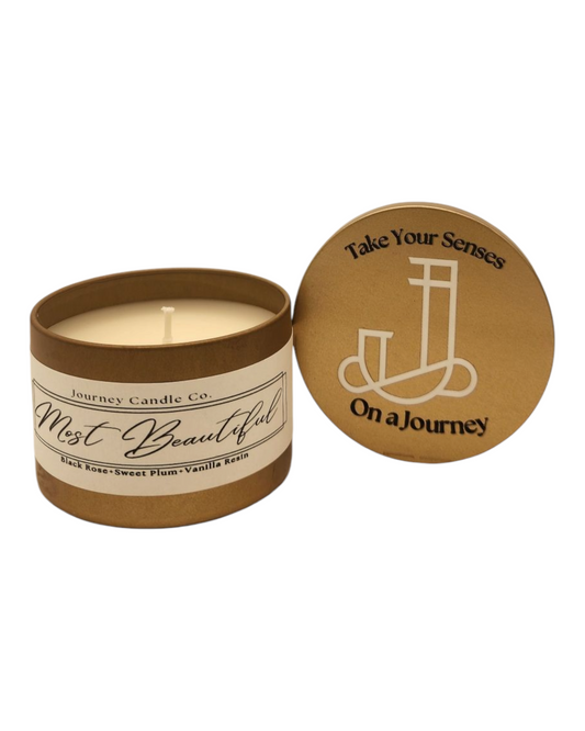 Most Beautiful 3oz gold candle tin with black rose, plum, and vanilla scent, paired with a complimentary branded satin travel gift bag.

