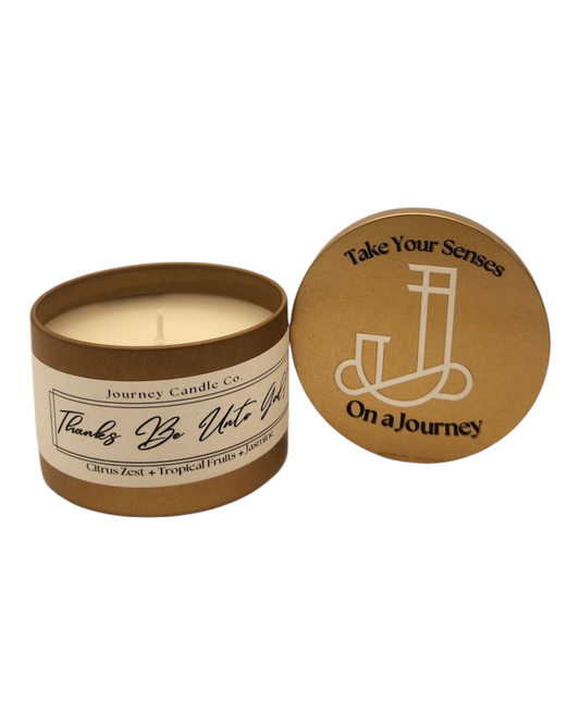 Thanks Be Unto God 3oz gold candle tin with tropical pineapple, goji berry, and driftwood scent, packaged in a complimentary branded satin travel bag.

